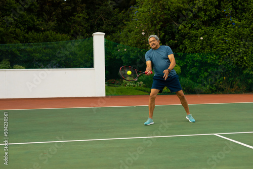 Full length of biracial senior man hitting ball with racket while playing tennis against trees © WavebreakMediaMicro