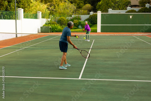 Full length of biracial senior man serving ball to senior woman while playing tennis in court