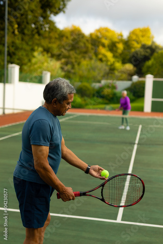 Biracial senior man serving tennis ball to senior wife while playing tennis in court against trees © WavebreakMediaMicro