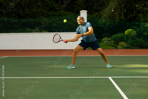 Full length of biracial senior man hitting tennis ball with racket while playing at tennis court © wavebreak3