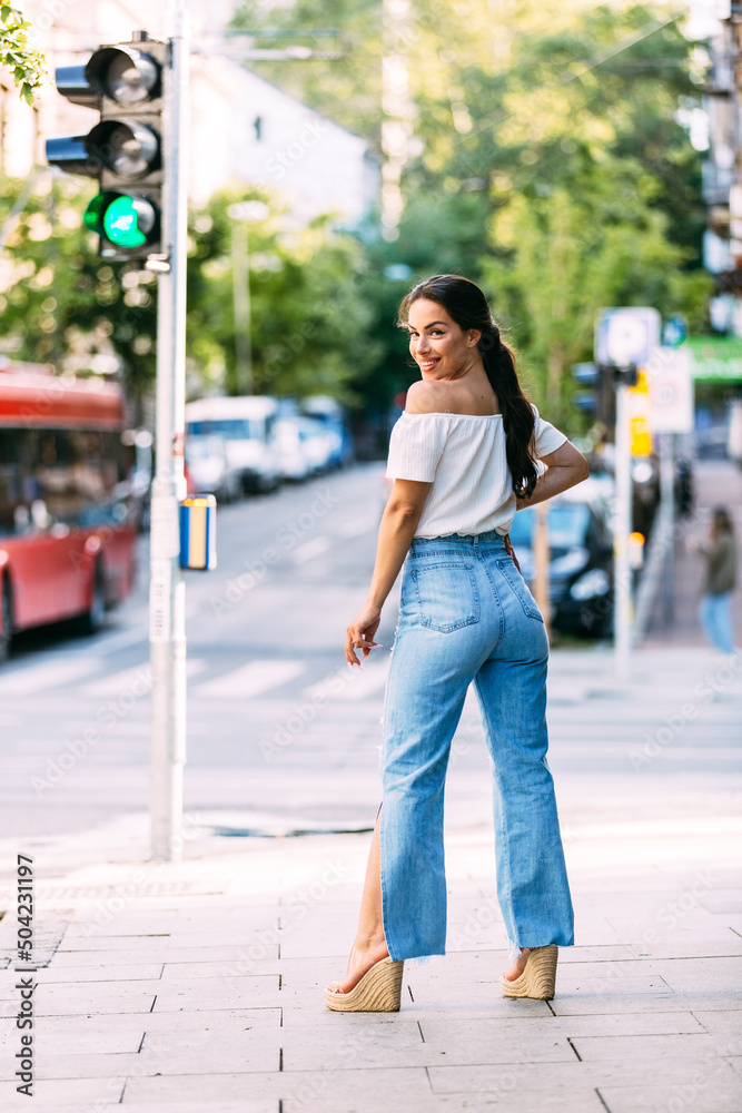 Full length portrait of beautiful female posing on street. Caucasian model dressed in casual jeans and shirt during fashion photo session