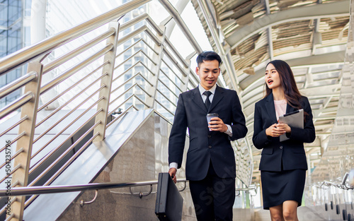 Two happy adult Asian businessman and woman or colleagues wearing formal suits, talking, walking together outdoor, going to work, holding cup of coffee with refreshment. Business, Lifestyle Concept.