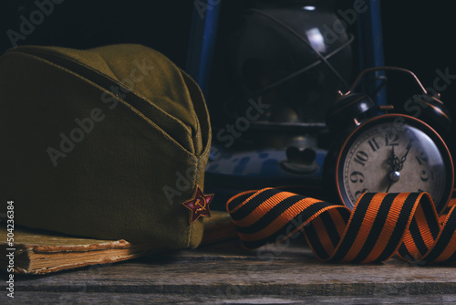 Russian soldiers cap,St. George ribbon, old book and an alarm clock with kerosene lamp. Victory Day is May 9th.