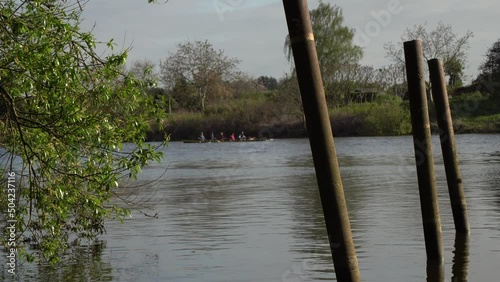 Rowers in rowboat rowing along Weser river close to Weserstadion under sunny blue sky. Some poles in foreground (parallax). Zoomed 4k pan left shot. (Bremen, Germany)