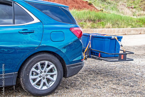 Blue hatchback car parked on gravel with hitch cargo carrier holding tied down rubber storage box on back - Closeup and cropped