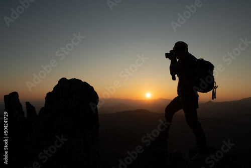 Silhouette of a travel photographer sitting on a rock and starting to shoot in the evening awaiting sunset at the peak of the national park. adventure experience concept