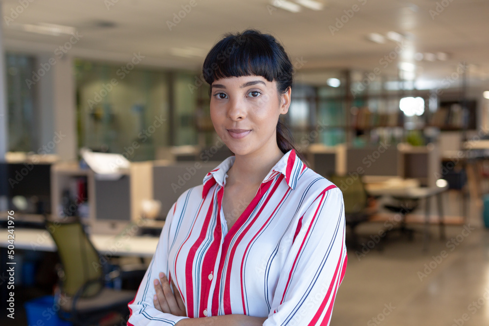 Fototapeta premium Portrait of confident young biracial businesswoman with arms crossed at modern workplace
