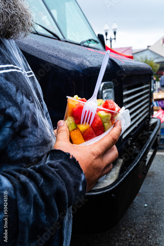 man holding cup of fruit pieces with white plastic fork in his hands purchased from street food vendor stall at fair
