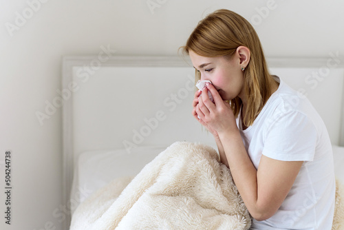 Sick young woman suffers from stuffy nose and sore throat. Upset sick Caucasian lady sitting in bed blowing her nose using a paper napkin. Cold and flu concept. A sick young woman coughs, sneezes