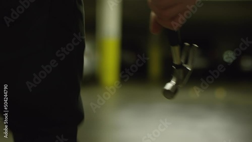 A man quickly aggressively walks through a car parking with a hammer in his hand. A tense scene of an upcoming crime. Thriller danger style. Character tracking effect. High quality closeup footage. 