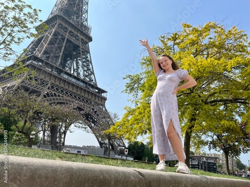 Canvas Print a cheerful teen girl stands against the backdrop of the Eiffel Tower in a beautiful dress, she raised her hand, you can advertise a travel agency trip to Paris France