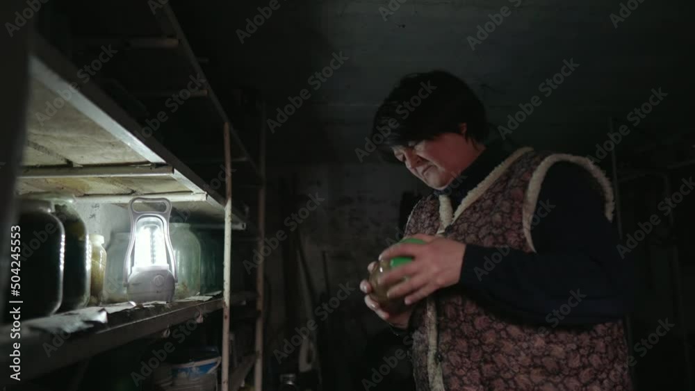 An elderly woman is sorting through jars of homemade pickled vegetables on the shelves in a dark old cellar. Typical rural life. Peasant woman in old clothes. Preparing for winter. An elderly woman is sorting through jars of homemade pickled vegetables on the shelves in a dark old cellar. Typical rural life. Peasant woman in old clothes. Preparing for winter.