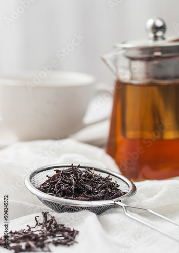 Strainer infusor with loose black tea on light cloth background with clear glass teapot and white ceramic cup