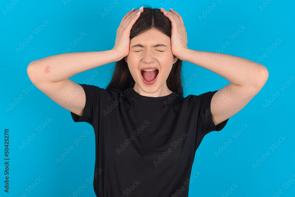 Shocked panic young caucasian woman wearing black T-shirt over blue background holding hands on head and screaming in despair and frustration. Shocked panic young caucasian woman wearing black T-shirt over blue background holding hands on head and screaming in despair and frustration.