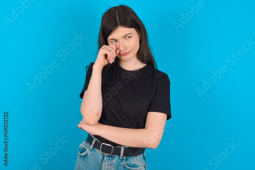 Unhappy young caucasian woman wearing black T-shirt over blue background crying while posing at camera whipping tears with hand. © Jihan