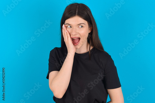 young caucasian woman wearing black T-shirt over blue background excited looking to the side hand on face. Advertisement and amazement concept.