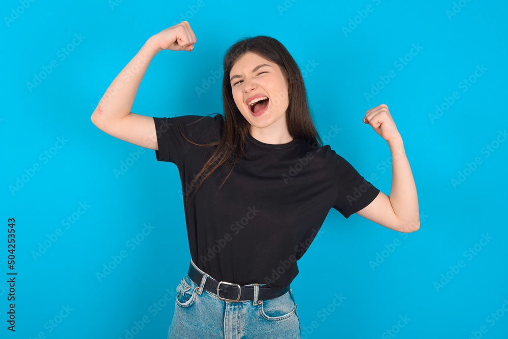 Yes I am winner. Portrait of charming delighted and excited young caucasian woman wearing black T-shirt over blue background raising up fist in triumph and victory smiling achieving success grinning Yes I am winner. Portrait of charming delighted and excited young caucasian woman wearing black T-shirt over blue background raising up fist in triumph and victory smiling achieving success grinning