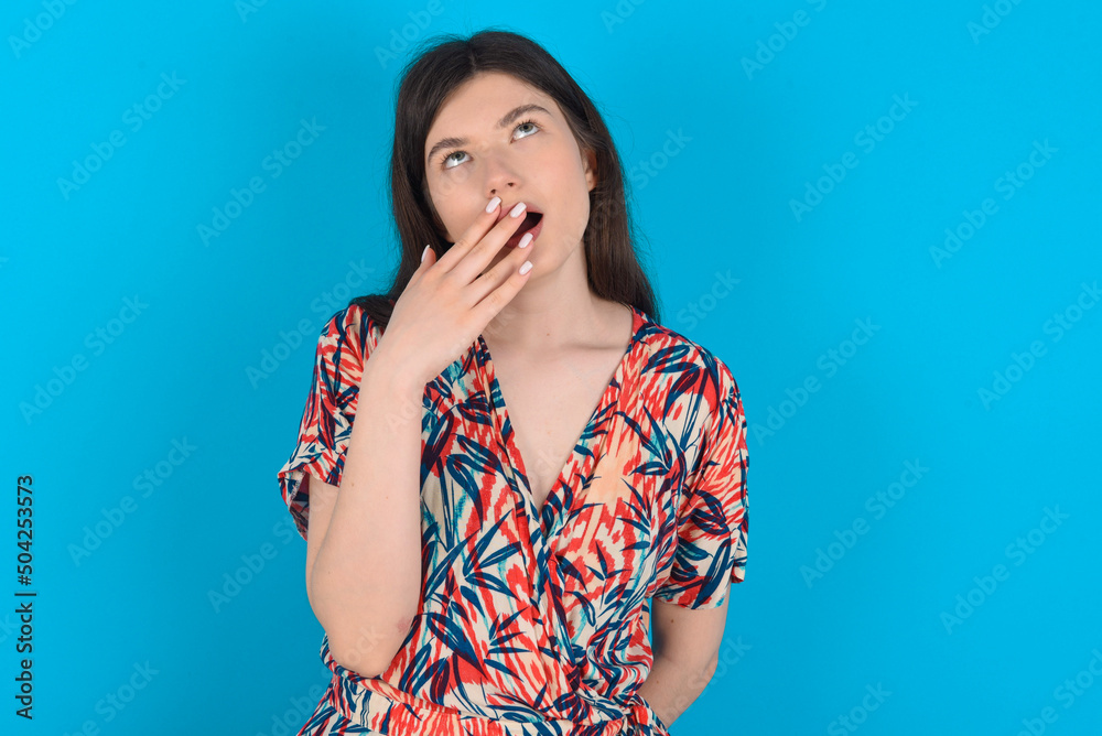 Sleepy young caucasian woman wearing floral dress over blue background yawning with messy hair, feeling tired after sleepless night, yawning, covering mouth with palm. Sleepy young caucasian woman wearing floral dress over blue background yawning with messy hair, feeling tired after sleepless night, yawning, covering mouth with palm.