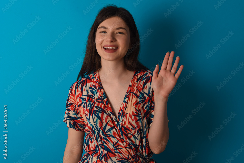 young caucasian woman wearing floral dress over blue background waiving saying hello or goodbye happy and smiling, friendly welcome gesture. young caucasian woman wearing floral dress over blue background waiving saying hello or goodbye happy and smiling, friendly welcome gesture.