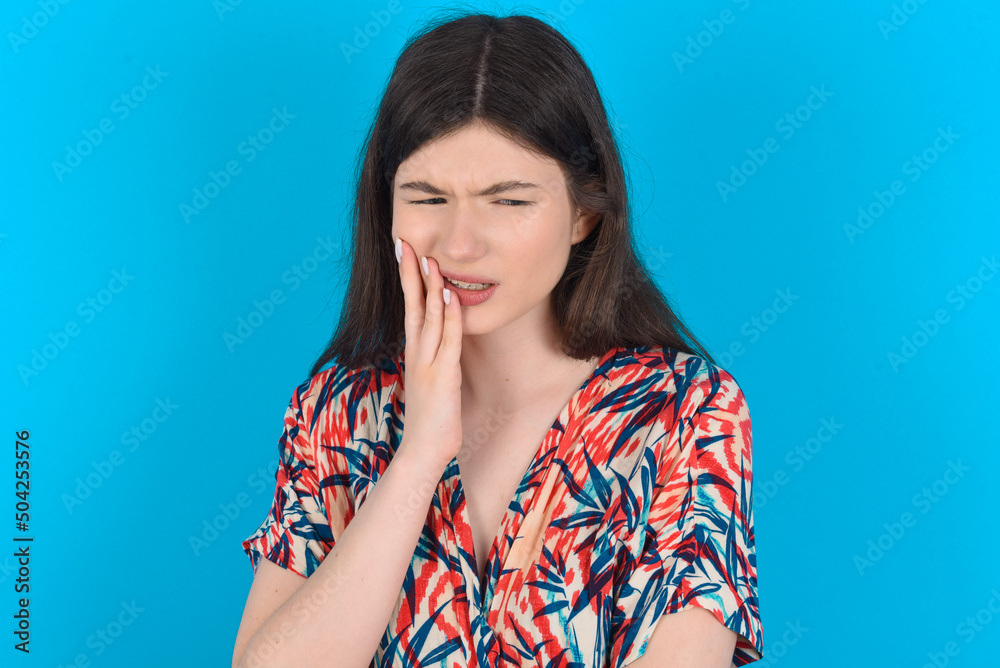 Tooth ache concept. young caucasian woman wearing floral dress over blue background feeling pain, holding his cheek with hand, suffering from bad toothache, looking at camera with painful expression Tooth ache concept. young caucasian woman wearing floral dress over blue background feeling pain, holding his cheek with hand, suffering from bad toothache, looking at camera with painful expression