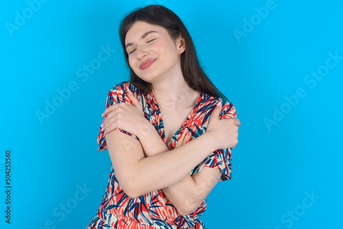 young caucasian woman wearing floral dress over blue background . Hugging oneself happy and positive, smiling confident. Self love and self care.