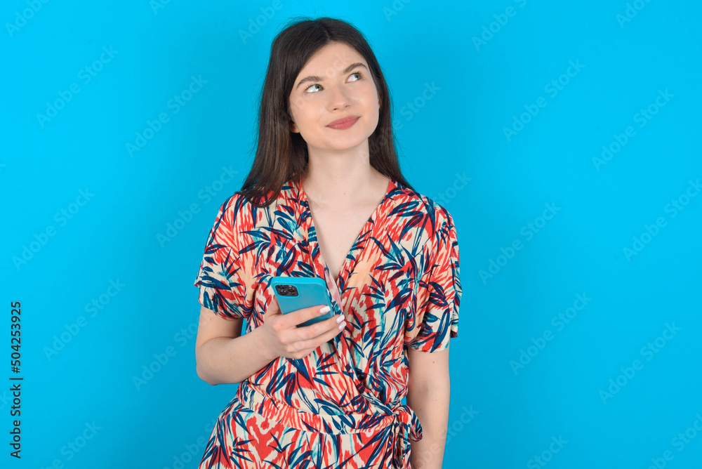 young caucasian woman wearing floral dress over blue background holds mobile phone uses high speed internet and social networks has online communication. Modern technologies concept young caucasian woman wearing floral dress over blue background holds mobile phone uses high speed internet and social networks has online communication. Modern technologies concept