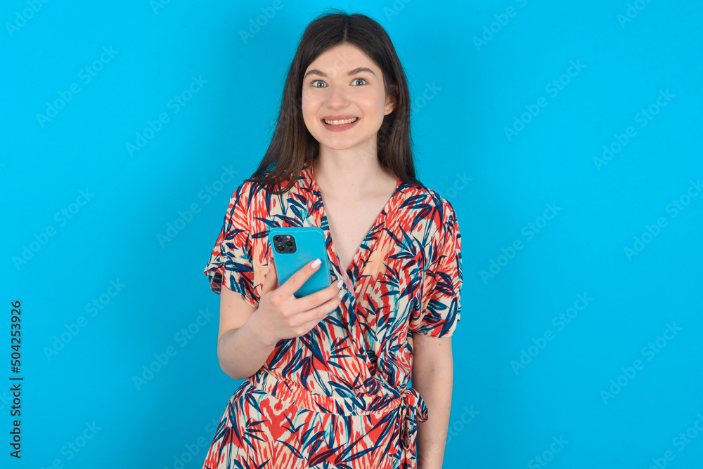 young caucasian woman wearing floral dress over blue background holds mobile phone in hands and rejoices positive news, uses modern cellular young caucasian woman wearing floral dress over blue background holds mobile phone in hands and rejoices positive news, uses modern cellular