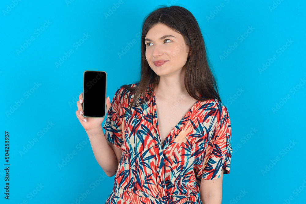 young caucasian woman wearing floral dress over blue background holds new mobile phone and looks mysterious aside shows blank display of modern cellular