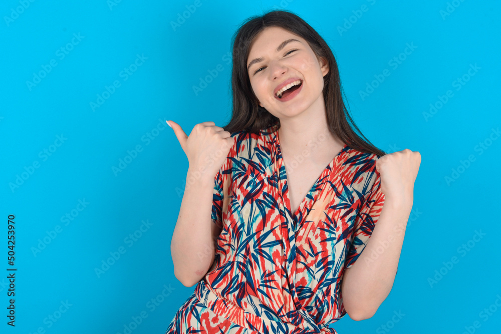 Fototapeta premium Hooray cool young caucasian woman wearing floral dress over blue background point back empty space hand fist