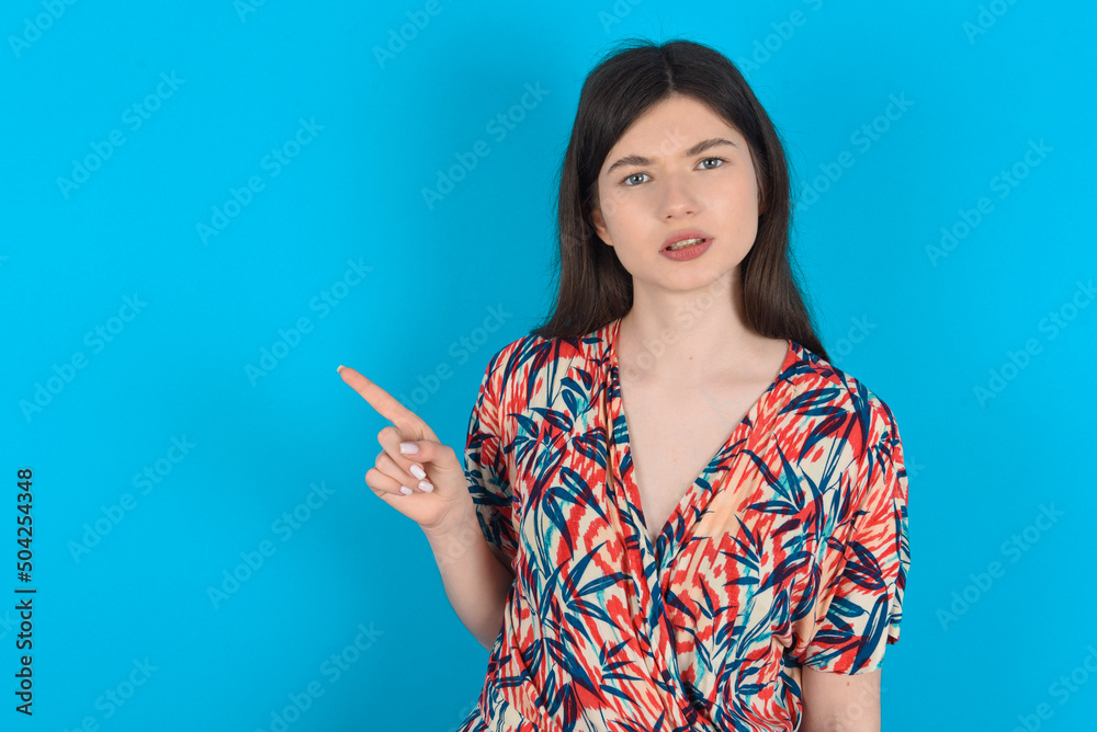 Serious young caucasian woman wearing floral dress over blue background smirks face points away on copy space shows something unpleasant. Look at this advertisement. Big price concept