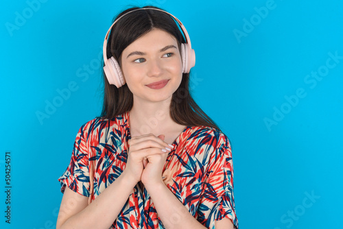 young caucasian woman wearing floral dress over blue background wears stereo headphones listening to music concentrated and looking aside with interest.