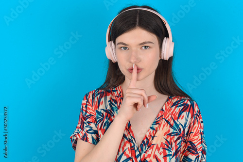 young caucasian woman wearing floral dress over blue background making hush gesture with finger on her lips wearing wireless headphones. Be quiet.