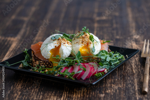 Bread toast, poached eggs, salmon, pea microgreens and fresh vegetables on plate in cafe, breakfast time