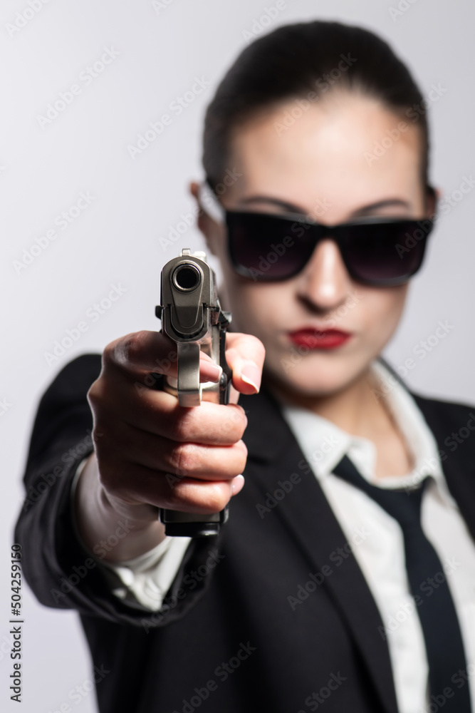 Beautiful and serious looking woman with sunglasses, classic black suit, tie and white shirt pointing pistol to camera. Model with red lipstick. Gun barrel is in camera focus and model is blurred Beautiful and serious looking woman with sunglasses, classic black suit, tie and white shirt pointing pistol to camera. Model with red lipstick. Gun barrel is in camera focus and model is blurred