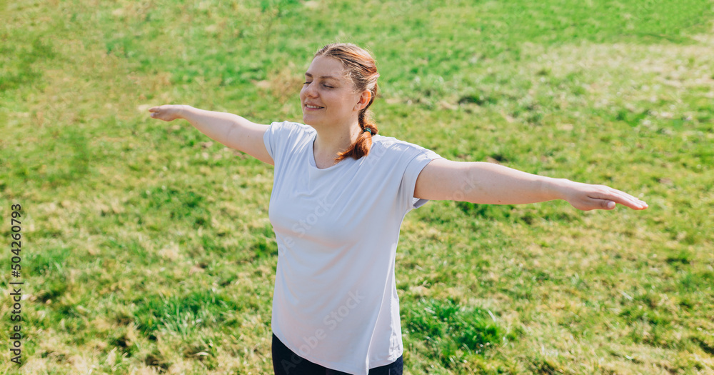 Redhead woman standing with arms outstretched closed eyes smiles feels love to life, welcoming new good day. Fitness female doing warmup exercise on nature background Redhead woman standing with arms outstretched closed eyes smiles feels love to life, welcoming new good day. Fitness female doing warmup exercise on nature background