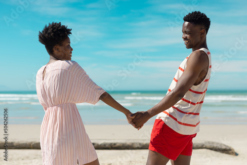 Happy african american young couple holding hands and looking at each other at beach against sky