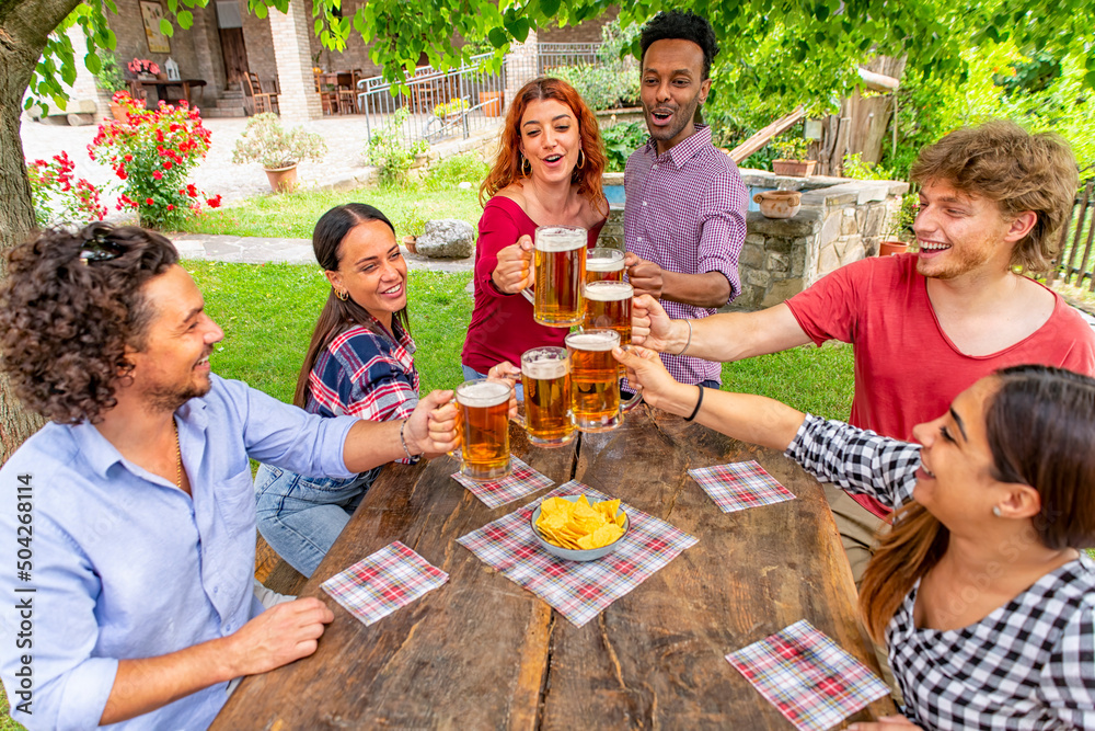 happy multiratial group of friends making a celebratory toast with beers on a picnic table outdoor in the garden. people gathering in a summer happy hour laughing in a party. joy and lifestyle concept