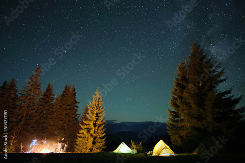 Bright illuminated tourist tents near glowing bonfire on camping site in dark mountain woods under night sky with sparkling stars. Active lifestyle and outdoor living concept