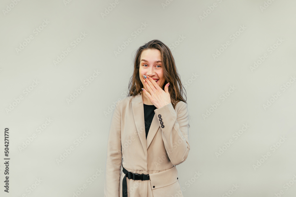 Beautiful woman in smart casual clothes looks to the side and smiles covering her mouth with her hand on a beige background, vertical. Beautiful woman in smart casual clothes looks to the side and smiles covering her mouth with her hand on a beige background, vertical.