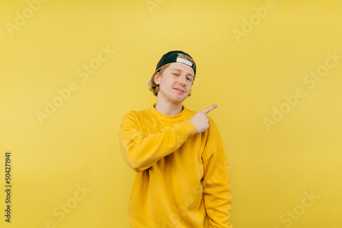 Happy guy in a cap and sweatshirt stands on a yellow background and points his finger to the side with a smile on his face.