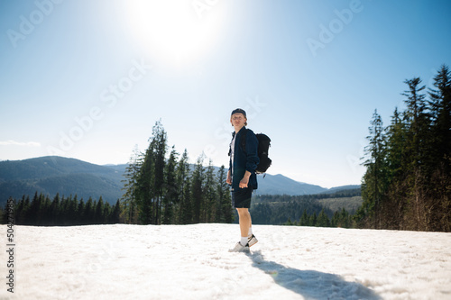 Handsome young man in light clothes stands in the snow in the mountains with a backpack on his back, posing for the camera while walking in a mountain resort in the spring.