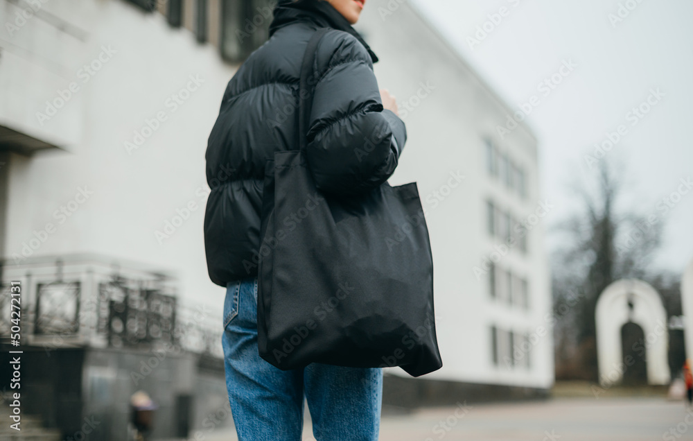 Close photo of a woman in a warm black jacket and with an eco bag in her hands walking down the street on a cloudy autumn day