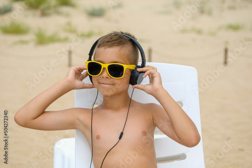 Young boy in sunglasses listens to music with headphones on the sea beach.