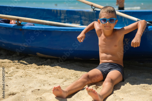 A young boy in a blue boat on the ocean. A child in sunglasses on the beach near the shore.
