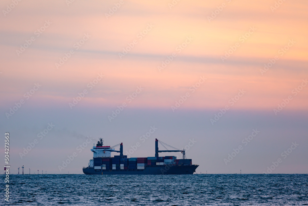 Large cargo ship with cranes carrying a load of shipping containers on the ocean at sunset.