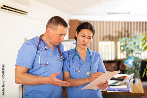 Portrait of two fellow doctors standing in medical office with papers in hands, focused on discussing of clinical diagnosis of patient