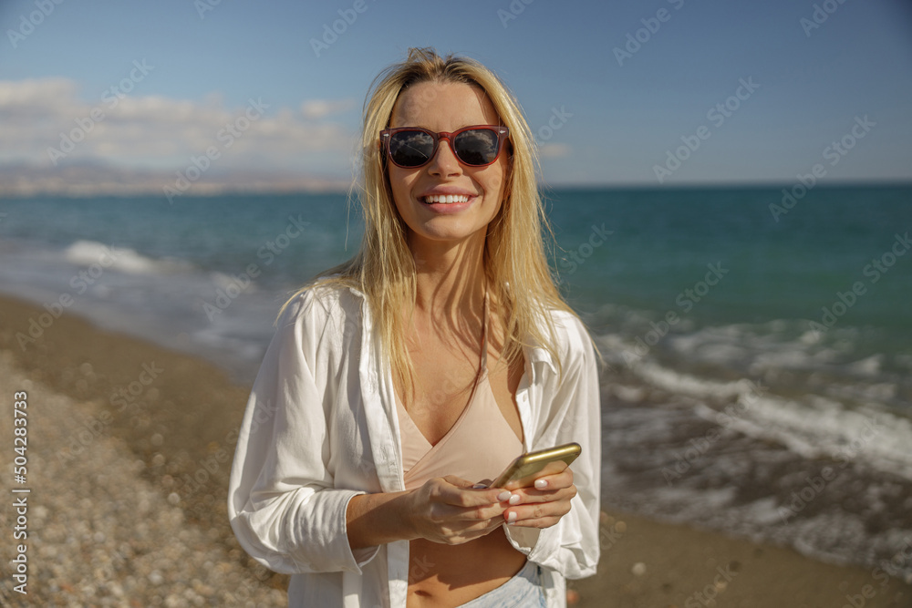 Portrait of happy lady in sunglasses using phone while standing on a beach