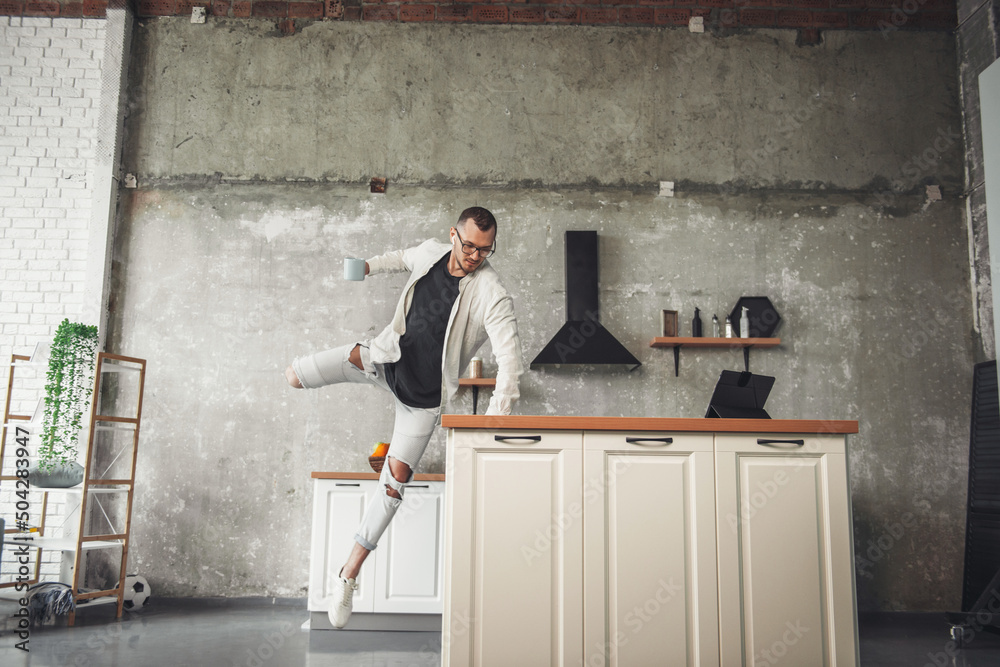 Front view of a man in jeans and a shirt wearing eyeglasses jumping in the kitchen holding a cup of coffee in his hand. Positive person. Front view.