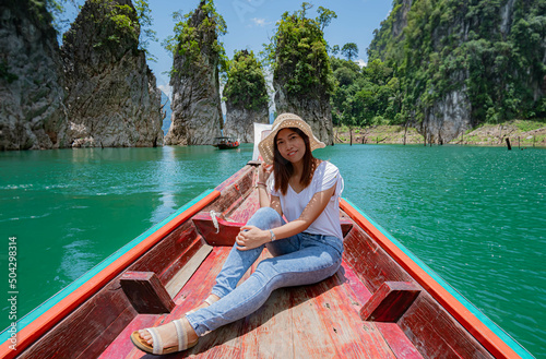 Happy young woman tourist with asian hat on the boat at lake and beautiful mountain view. Woman on Boat in landmark at Ratchaprapha Dam Khao Sok National Park in Thailand.