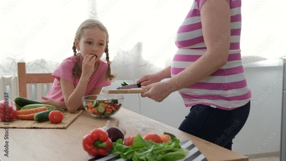 Little Girl Eating Vegetables while her Pregnant Mother Cooking Salad in the Kitchen. Healthy Food and Maternity Concept Little Girl Eating Vegetables while her Pregnant Mother Cooking Salad in the Kitchen. Healthy Food and Maternity Concept